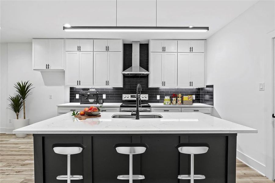 Kitchen featuring light stone counters, a center island with sink, white cabinets, and wall chimney range hood