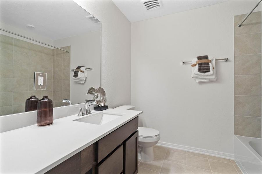 Secondary bath features tile flooring, bath/shower combo with tile surround, stained wood cabinets, beautiful light countertops, mirror, dark, sleek fixtures and modern finishes.