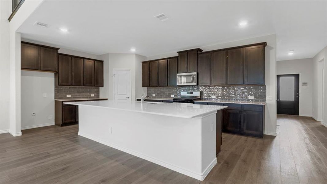 Spacious kitchen featuring a large white island with a light-toned countertop, dark wood-finish cabinetry, a subway tile backsplash, and stainless steel appliances