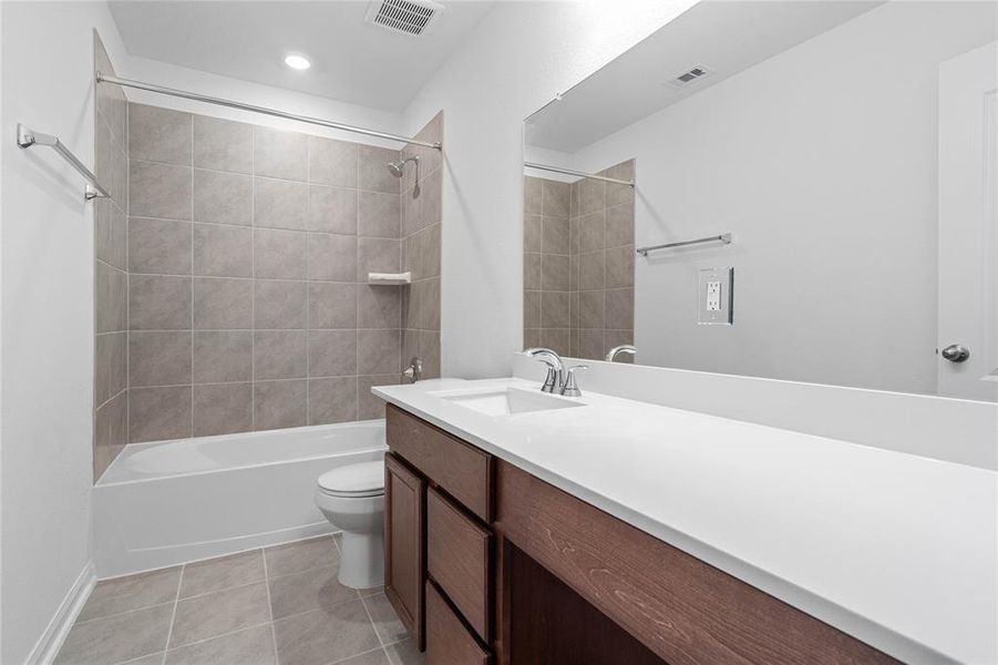 Secondary bath features tile flooring, bath/shower combo with tile surround, wood cabinets, beautiful light countertops, mirror, dark, sleek fixtures and modern finishes.