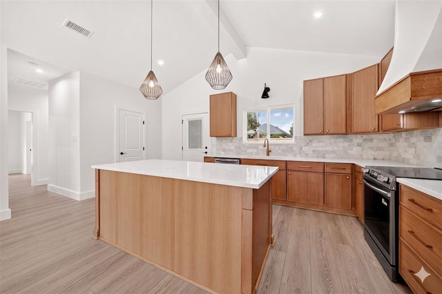 Spacious kitchen featuring a large central island with a white solid surface countertop, light wood cabinetry, white vaulted ceilings, and wood-finish flooring