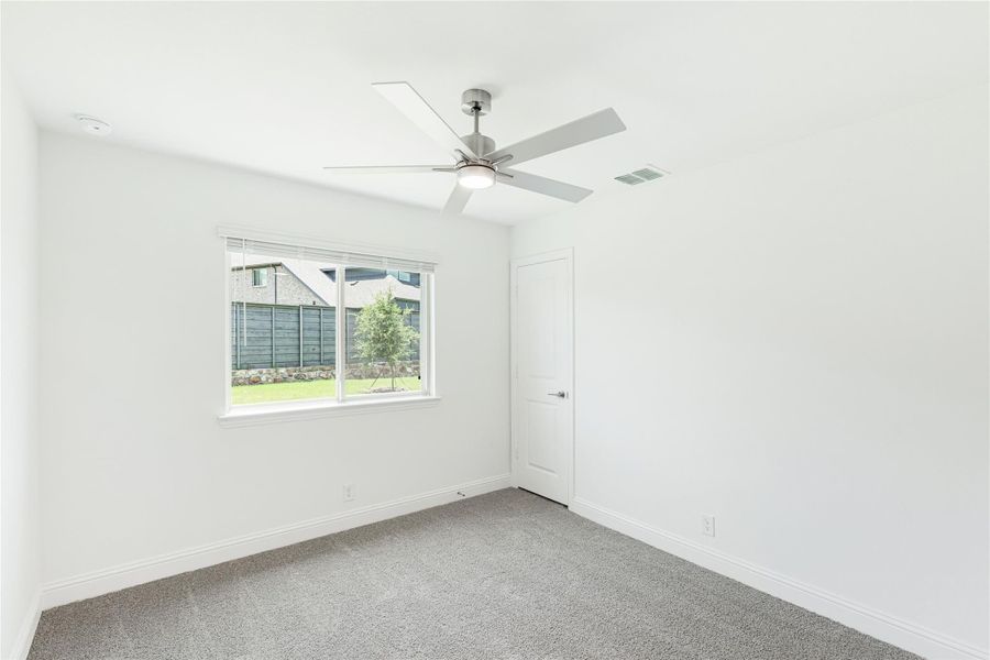 Empty bedroom with gray carpet, ceiling fan, white walls, and a window overlooking a backyard Empty bedroom with gray carpet, ceiling fan, white walls, and a window overlooking a backyard