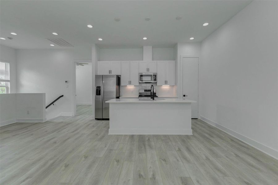 Modern kitchen and living area centered around a sleek white island with quartz-style counters, stainless appliances, black faucet, white cabinetry, and under-cabinet lighting. The open plan feels bright and refined, offering a stylish hub for cooking, casual dining, entertaining, and everyday connection.