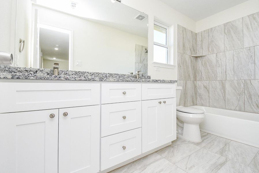 Modern bathroom with white vanity, granite countertop, large mirror, tiled walls, and a bathtub with a shower area. Bright and clean design.