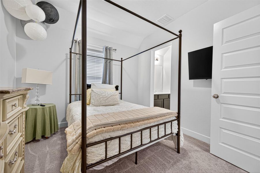 Cozy bedroom with a four-poster bed, neutral tones, and carpeted floor. Features a dresser, round side table, wall-mounted TV, and an ensuite sink area. Natural light filters through a window with curtains. Cozy bedroom with a four-poster bed, neutral tones, and carpeted floor. Features a dresser, round side table, wall-mounted TV, and an ensuite sink area. Natural light filters through a window with curtains.