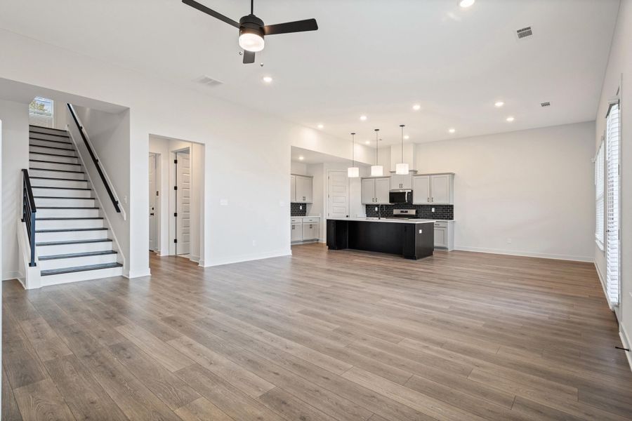 Open-concept living space featuring wood-finish flooring, a modern kitchen with a large island and pendant lighting, and a staircase with a black handrail Open-concept living space featuring wood-finish flooring, a modern kitchen with a large island and pendant lighting, and a staircase with a black handrail