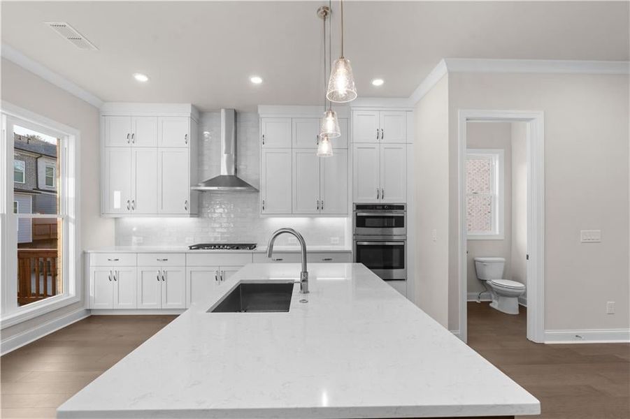 Kitchen with pendant lighting, ornamental molding, white cabinetry, light stone counters, and a kitchen island with sink