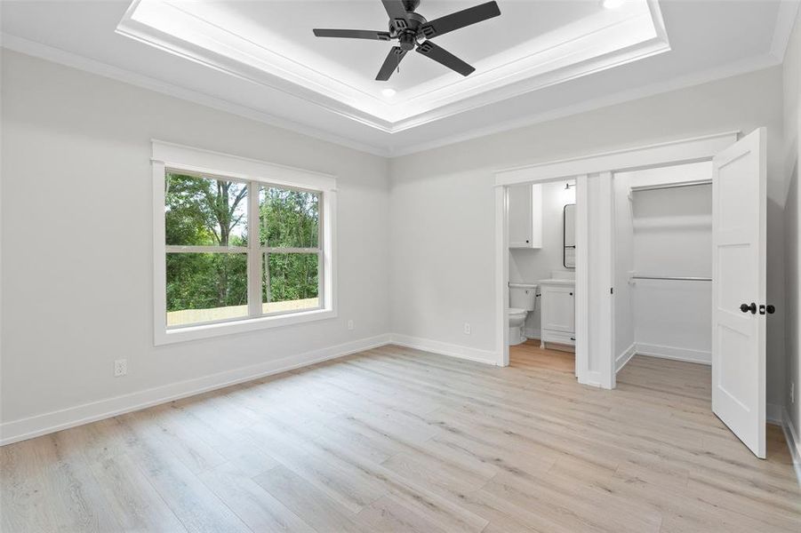 Unfurnished bedroom with a raised ceiling, crown molding, light wood finished floors, connected bathroom, and a walk in closet