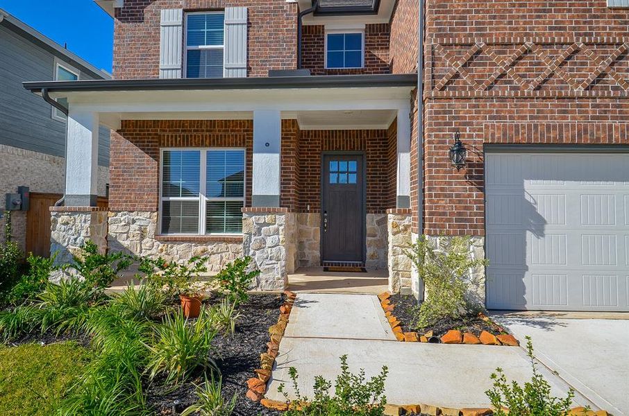 This home features a charming brick and stone exterior with a welcoming front porch. It has a modern front door, well-maintained landscaping, and a spacious driveway leading to a garage.