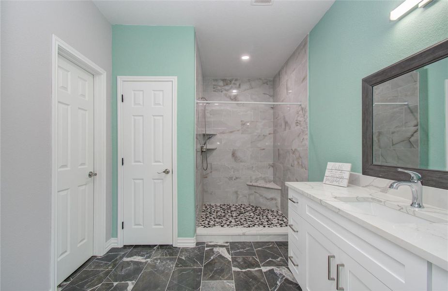 This bathroom features a modern design with a spacious walk-in shower adorned with elegant gray tiles. It includes a sleek vanity with a large mirror and ample storage. The room is accented with a calming teal wall and contemporary lighting.