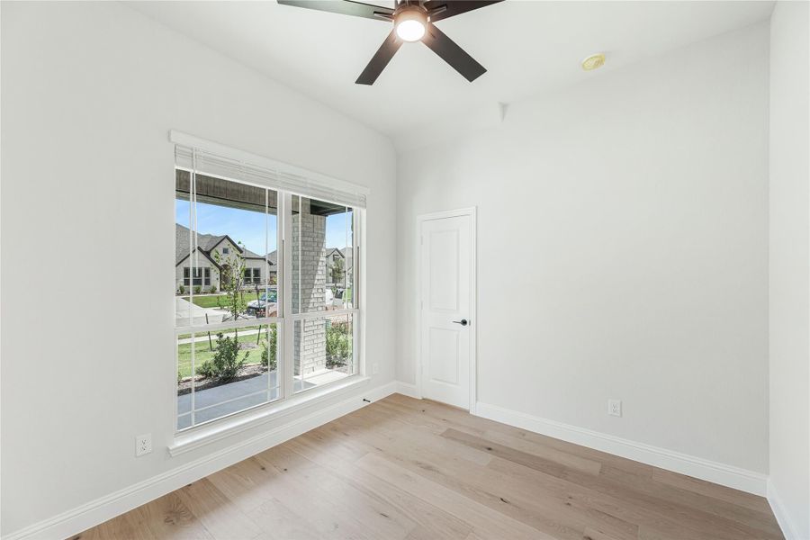 Empty bedroom with light wood floors, ceiling fan, and large window overlooking neighborhood