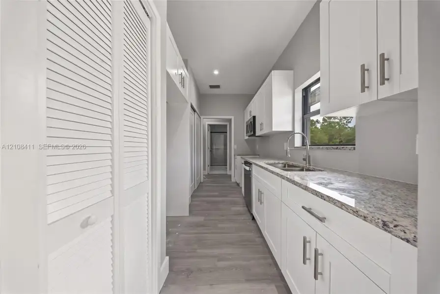 Galley kitchen featuring white Shaker-style cabinetry, stone-finish countertops, stainless steel undermount sink with a gooseneck faucet, wood-finish flooring, and white louvered-door closets