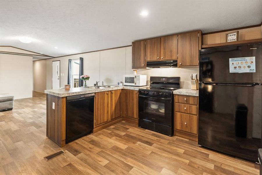 Full kitchen featuring wood-finish flooring, wood-look cabinetry, black appliances, a double-basin sink, and light-toned countertops Full kitchen featuring wood-finish flooring, wood-look cabinetry, black appliances, a double-basin sink, and light-toned countertops