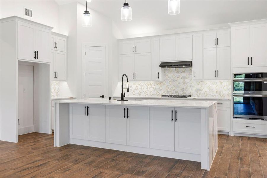 Spacious kitchen featuring white cabinetry, a large center island with a stone countertop, a decorative tile backsplash, and wood-finish flooring Spacious kitchen featuring white cabinetry, a large center island with a stone countertop, a decorative tile backsplash, and wood-finish flooring