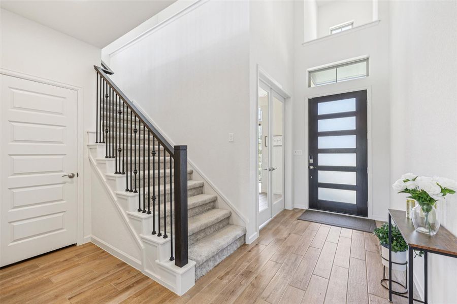 The entryway features light-colored wood-look flooring, a carpeted staircase with dark iron balusters, and a modern dark-framed front door with frosted glass panels