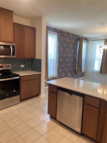 Kitchen with stainless steel appliances, brown cabinets, light tile patterned floors, decorative backsplash, and light stone counters Kitchen with stainless steel appliances, brown cabinets, light tile patterned floors, decorative backsplash, and light stone counters