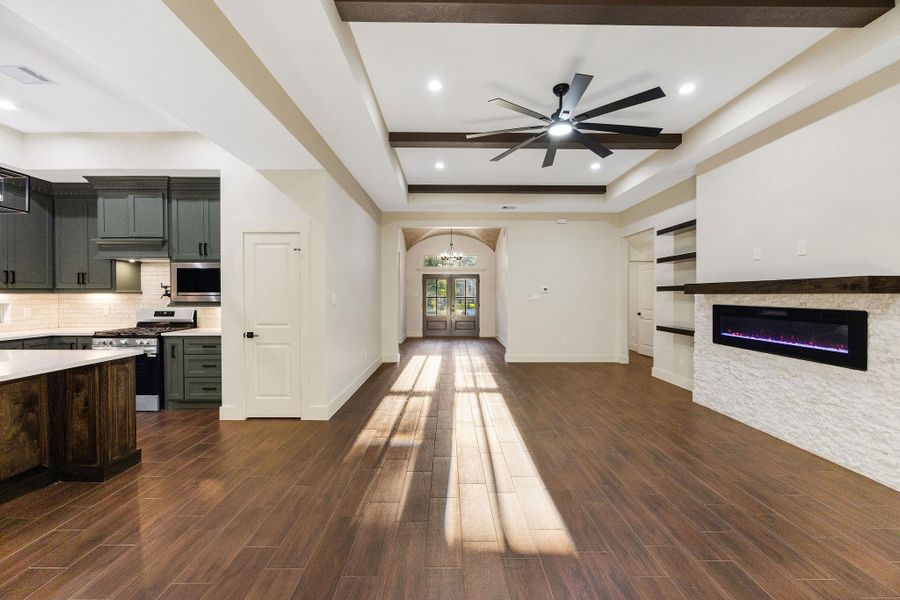 This spacious open-concept living area with hardwood floors, a modern kitchen featuring dark cabinets and stainless steel appliances, a large ceiling fan, and a sleek electric fireplace set into a white stone accent wall. Natural light streams in from the entrance, creating a warm and inviting atmosphere.