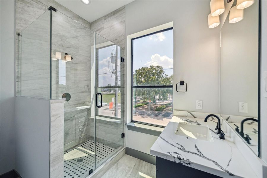 Bright and inviting, this bathroom showcases a walk-in shower with frameless glass and tile surround, paired with a vanity topped in quartz with clean-lined fixtures. A large window allows natural light to fill the space while offering a view toward the front of the property.