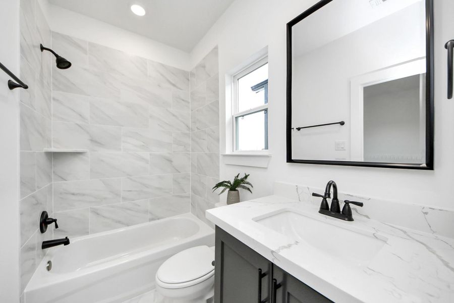 This modern bathroom features a sleek design with marble-patterned tiles, a built-in bathtub, and a stylish vanity with a black-framed mirror. The room is brightened by natural light from a window, and includes black fixtures and fittings for a contemporary look. This modern bathroom features a sleek design with marble-patterned tiles, a built-in bathtub, and a stylish vanity with a black-framed mirror. The room is brightened by natural light from a window, and includes black fixtures and fittings for a contemporary look.