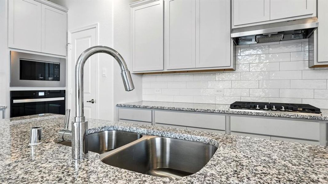 Kitchen featuring a double basin stainless steel sink with a chrome gooseneck faucet, granite countertops, white subway tile backsplash, white cabinetry, and stainless steel appliances including a built-in oven and microwave