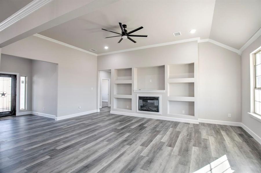 Unfurnished living room with plenty of natural light, built in shelves, crown molding, a glass covered fireplace, and ceiling fan