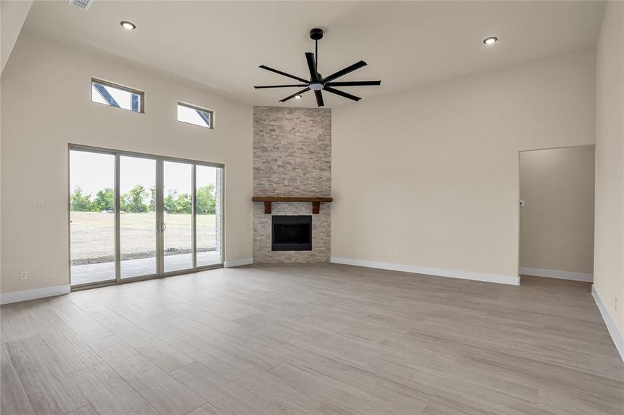 Unfurnished living room with ceiling fan, a towering ceiling, light wood-style flooring, a stone fireplace, and recessed lighting Unfurnished living room with ceiling fan, a towering ceiling, light wood-style flooring, a stone fireplace, and recessed lighting