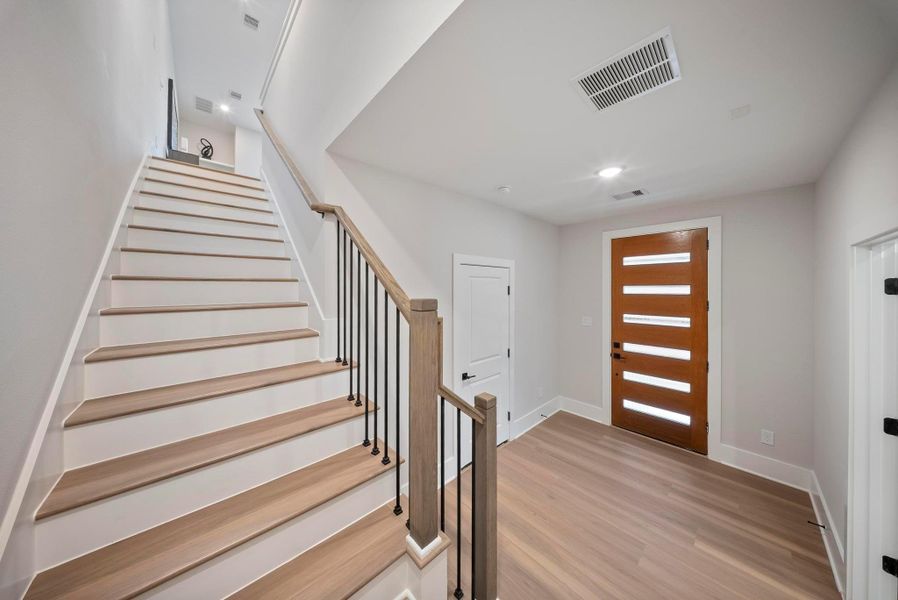 Bright and modern entryway featuring a sleek wooden staircase with black railings, contemporary front door with horizontal glass panels, and light wood flooring.