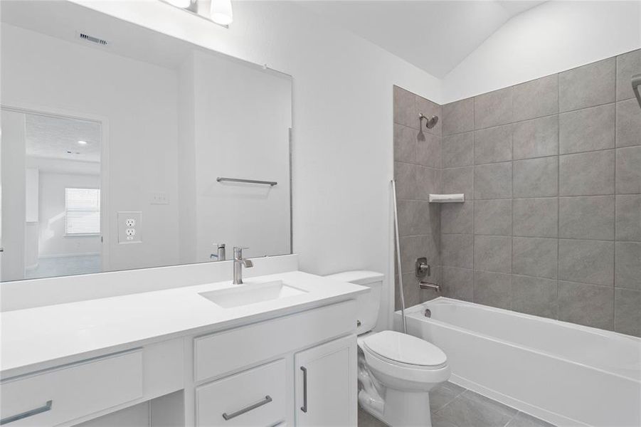 Secondary bath features tile flooring, bath/shower combo with tile surround, stained wood cabinets, beautiful light countertops, mirror, dark, sleek fixtures and modern finishes.