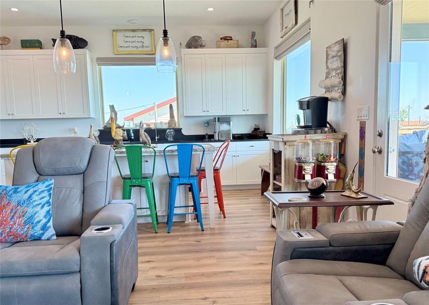 This cozy living space features modern touches with light wood flooring, comfortable recliners, and a vibrant kitchen area. The kitchen includes white cabinetry, a breakfast bar with colorful metal stools, and pendant lighting. Large windows provide plenty of natural light, enhancing the inviting atmosphere.