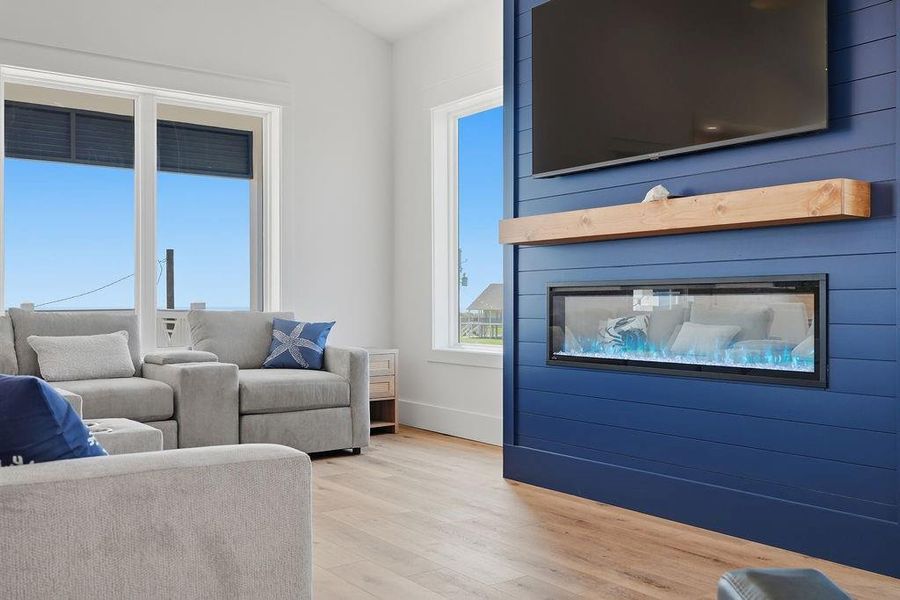 This modern living room features a cozy gray sectional, large windows for natural light, and a stylish blue accent wall with a sleek electric fireplace and mounted TV. The light wood flooring adds warmth to the space.