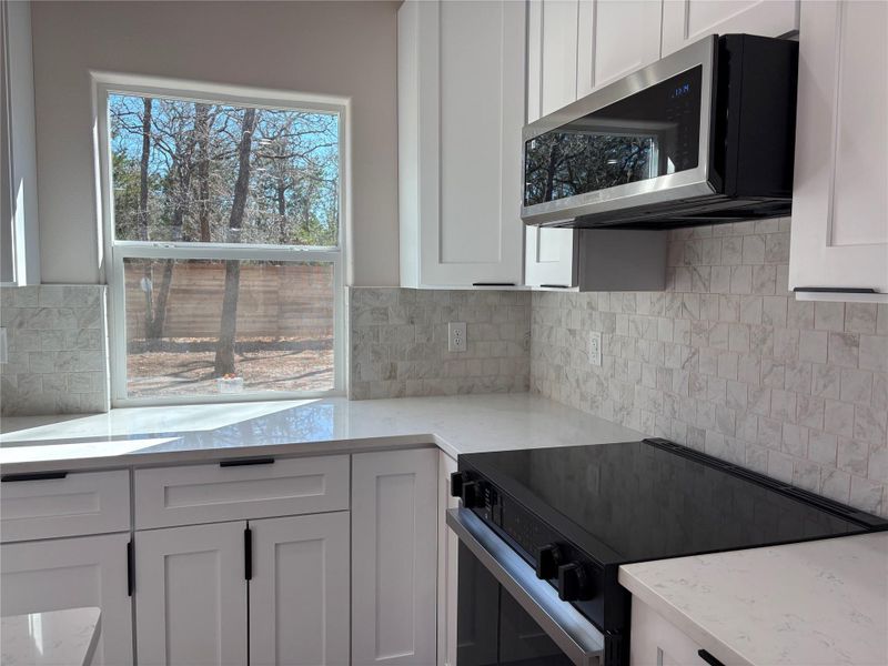 Kitchen with electric range, light stone countertops, stainless steel microwave, white cabinetry, and backsplash