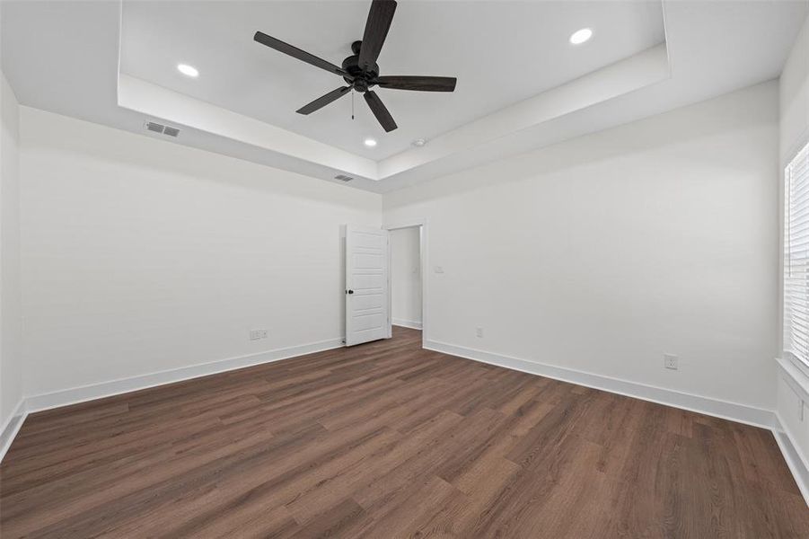 This spacious room features wood plank flooring, white walls, and a tray ceiling with recessed lighting and a ceiling fan