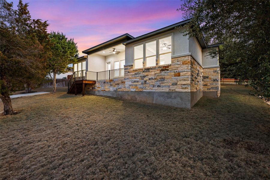 Back of property at dusk featuring stone siding, a lawn, ceiling fan, and stucco siding