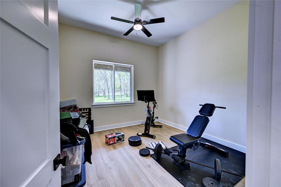 This room is set up as a home gym with exercise equipment, light wood flooring, and a ceiling fan. A window provides natural light and a view of the outdoors.