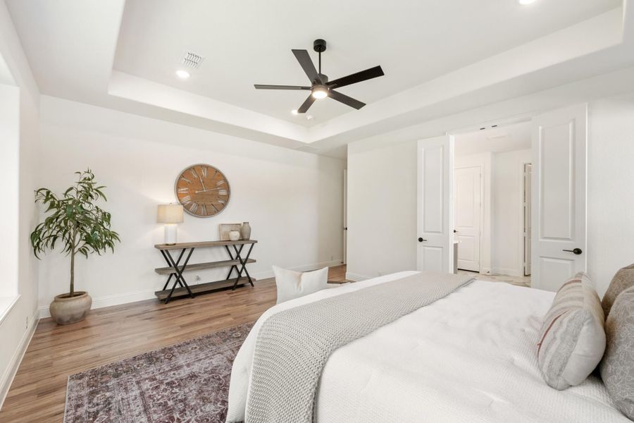 Bedroom with tray ceiling, ceiling fan, hardwood floors, and white walls with a console table and potted plant