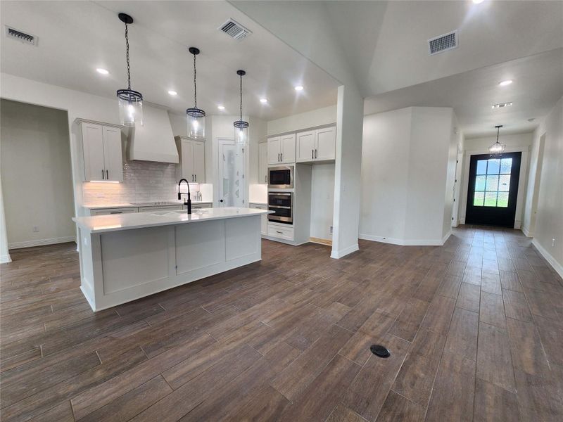 Open-concept kitchen featuring a large central island with a solid surface countertop, white cabinetry, a subway tile backsplash, and built-in double ovens
