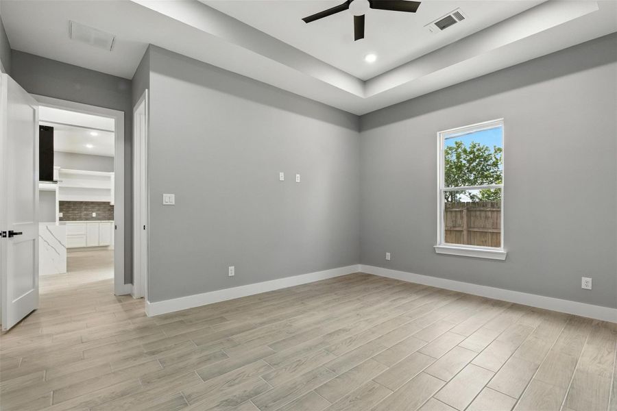 A spacious primary room with light flooring and gray walls, featuring a tray ceiling with recessed lighting and a ceiling fan. There's a large window providing natural light and a view outside. The room connects to another area through an open door, revealing a glimpse of a modern kitchen or living space.