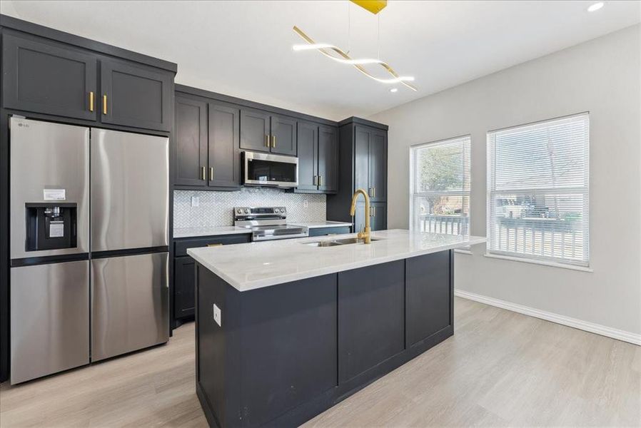 Kitchen with stainless steel appliances, decorative backsplash, light wood-style floors, a kitchen island with sink, and a sink