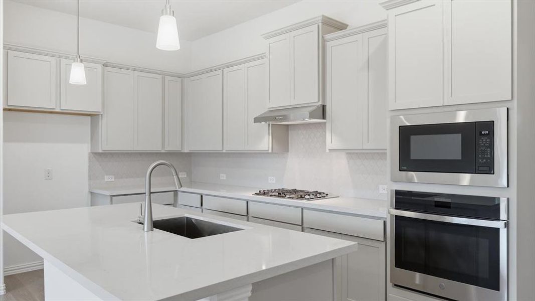 Modern kitchen featuring light gray cabinetry, built-in stainless steel appliances, a center island with an undermount sink, pendant lighting, and a neutral tile backsplash Modern kitchen featuring light gray cabinetry, built-in stainless steel appliances, a center island with an undermount sink, pendant lighting, and a neutral tile backsplash