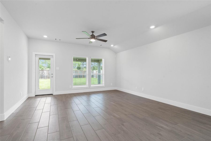 A bright and airy open-concept space featuring tile flooring, sloped ceilings with recessed lighting and ceiling fan, abundant natural light from large windows (fitted with blinds), and an access door to the back covered patio.