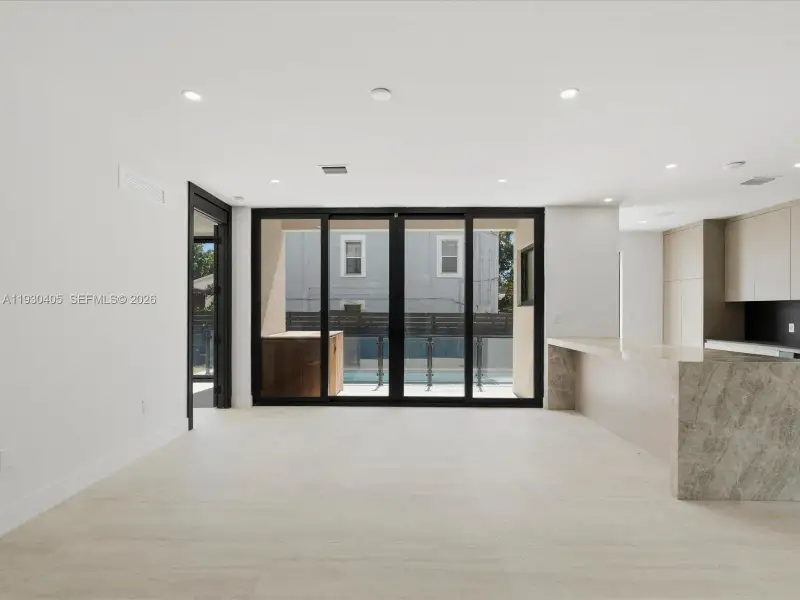 Open-concept interior featuring large-format stone tile flooring, a stone-top kitchen island with waterfall edge, flat-panel cabinetry, and multi-panel sliding glass doors Open-concept interior featuring large-format stone tile flooring, a stone-top kitchen island with waterfall edge, flat-panel cabinetry, and multi-panel sliding glass doors
