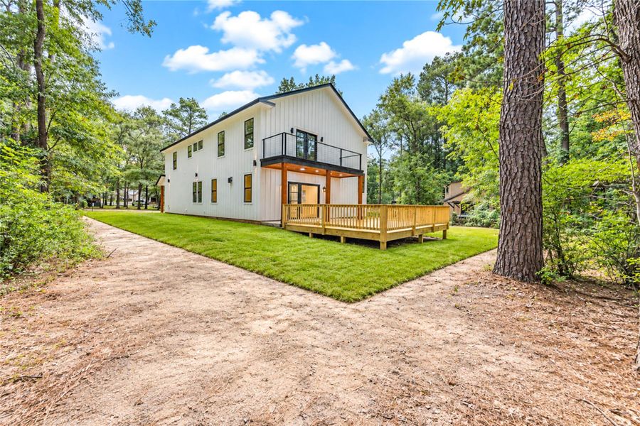 Modern two-story home with a spacious wooden deck, nestled in a serene wooded setting with lush greenery and a well-maintained lawn.