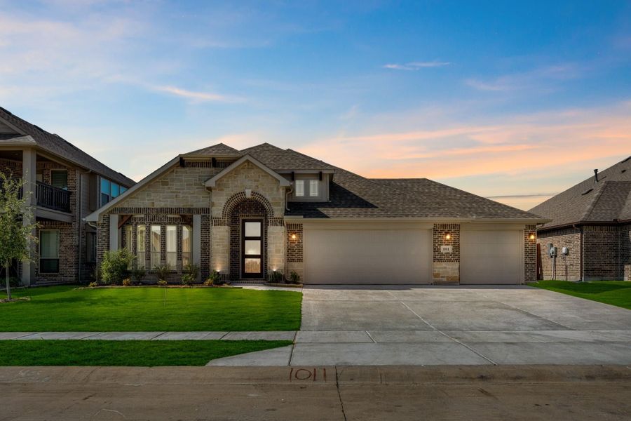 Single-story brick and stone home with arched entry, three-car garage, and green front lawn at dusk Single-story brick and stone home with arched entry, three-car garage, and green front lawn at dusk