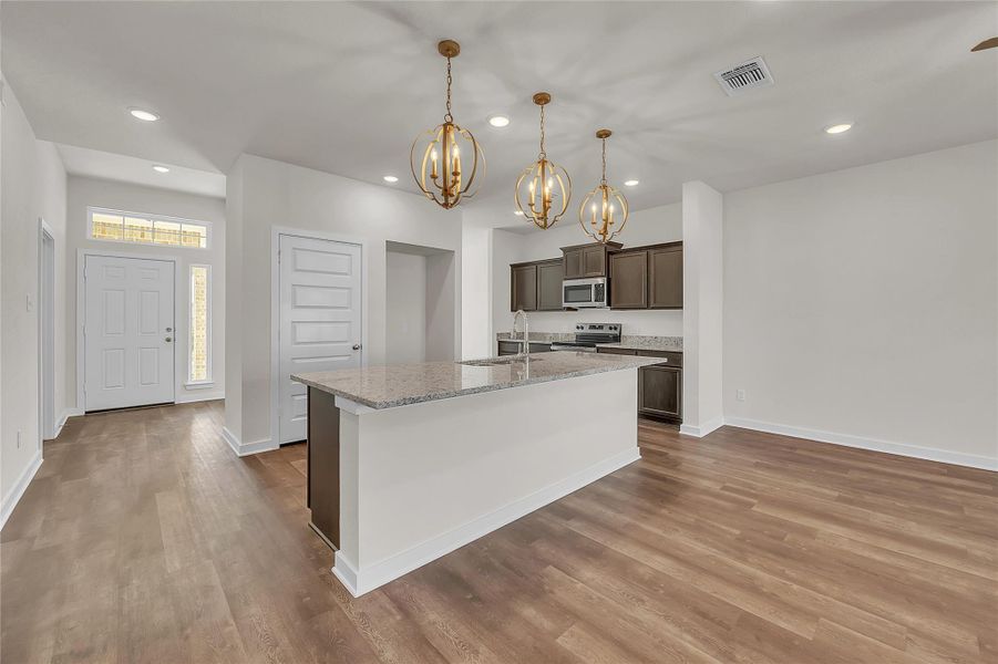Kitchen with dark brown cabinets, light stone counters, decorative light fixtures, stainless steel appliances, and light wood-style floors Kitchen with dark brown cabinets, light stone counters, decorative light fixtures, stainless steel appliances, and light wood-style floors