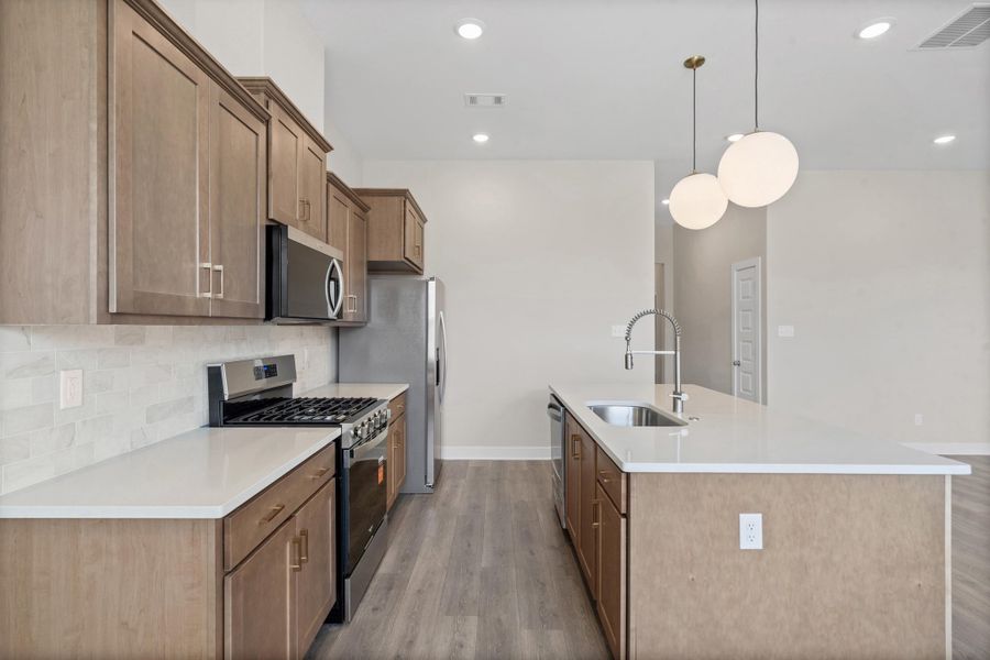 Kitchen featuring light wood-finish cabinetry, stainless steel appliances, a white tile backsplash, and an island with a stainless steel sink and modern pendant lighting