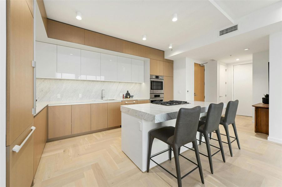 A modern, open-concept kitchen, dining and living area. The kitchen features a sleek, minimalist design with a large island at its center, topped with white-colored stone countertop. The island includes a built-in gas cooktop and stainless steel oven. Custom cabinetry seamlessly conceals the integrated refrigerator, maintaining clean lines and a refined aesthetic.