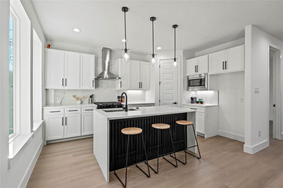 Kitchen featuring white cabinetry, a kitchen island with sink, stainless steel microwave, and wall chimney range hood