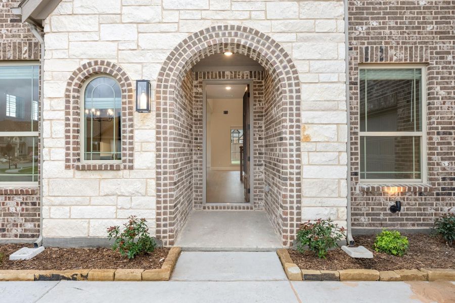 Home entrance with brick arched doorway, stone exterior, wall sconce, and small landscaped shrubs along walkway Home entrance with brick arched doorway, stone exterior, wall sconce, and small landscaped shrubs along walkway