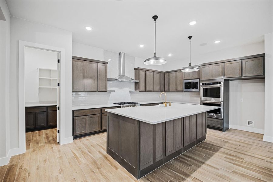 Kitchen with dark brown cabinets, wall chimney range hood, light countertops, light wood-style flooring, and appliances with stainless steel finishes Kitchen with dark brown cabinets, wall chimney range hood, light countertops, light wood-style flooring, and appliances with stainless steel finishes