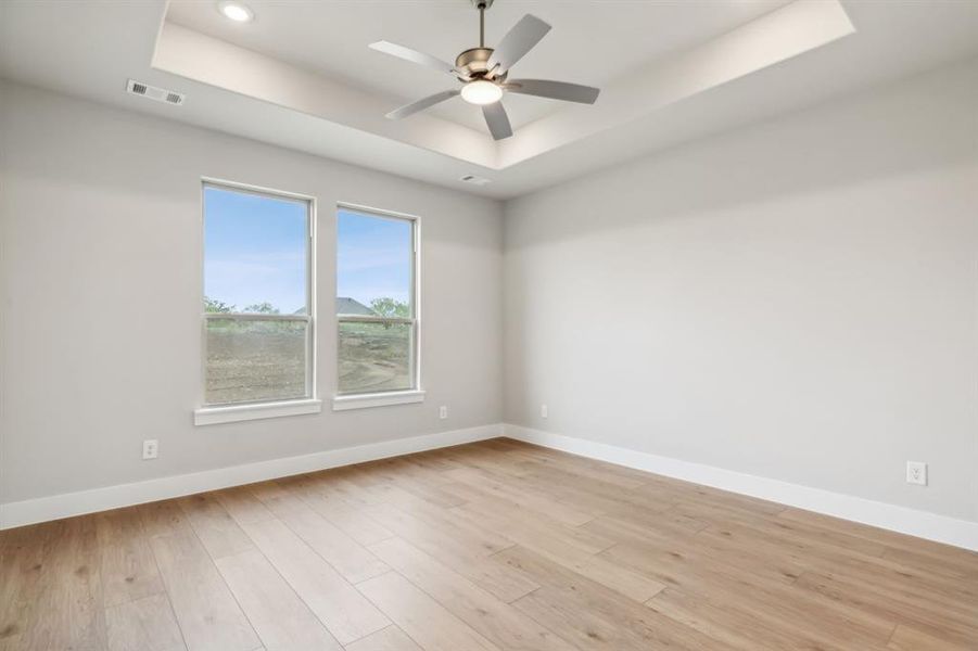 Empty room with ceiling fan, light wood-type flooring, and a tray ceiling